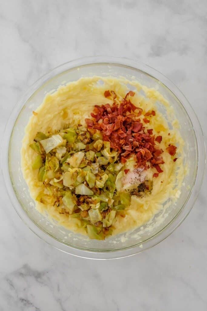 A glass bowl containing mashed potatoes topped with cooked cabbage mixture, chopped bacon, and seasonings.