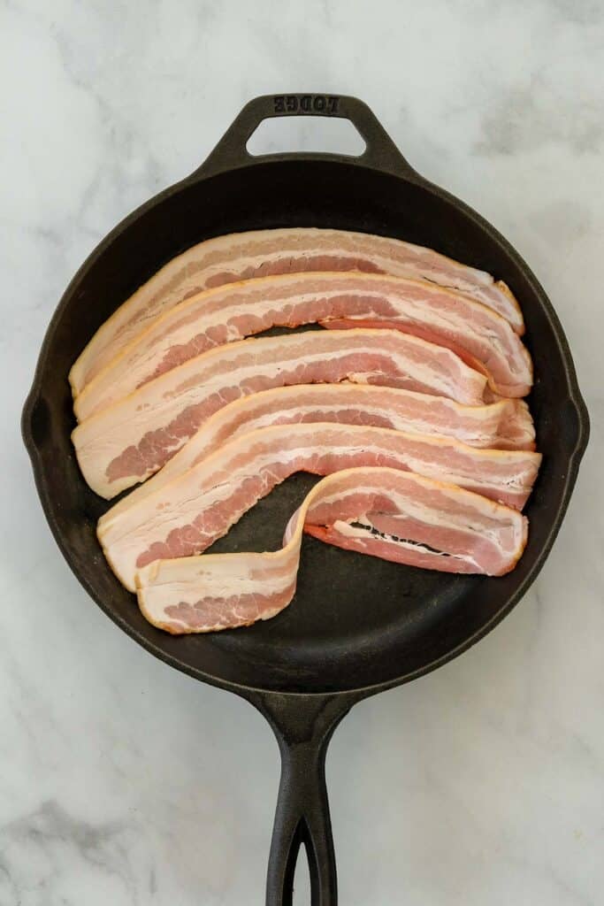 Raw strips of bacon are arranged in a single layer inside a black cast iron skillet.