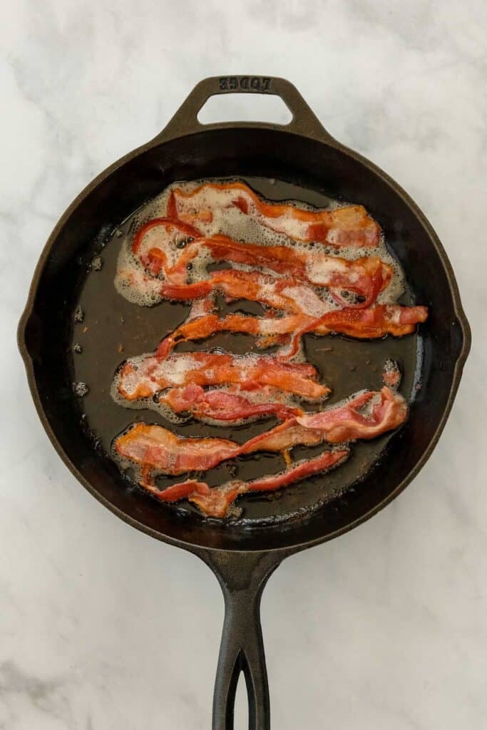 Bacon strips cooking in a cast iron skillet.