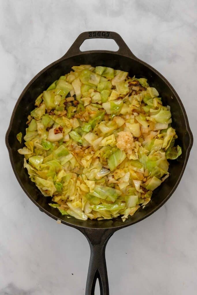 A cast iron skillet filled with sautéed chopped cabbage, onions and minced garlic on a light marble surface.