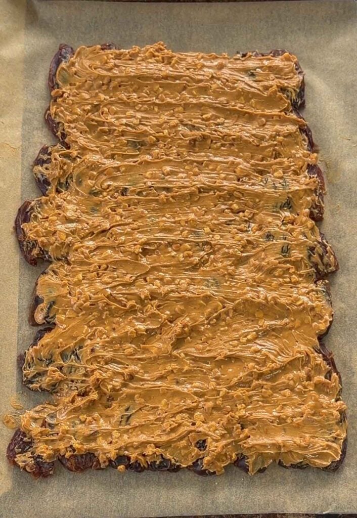 A sheet of dates topped with a thick layer of chunky peanut butter, placed on parchment paper.