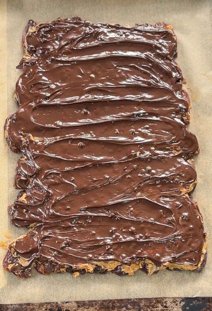 A rectangular sheet of dates topped with a thick layer of glossy melted chocolate and peanut butter, placed on parchment paper.