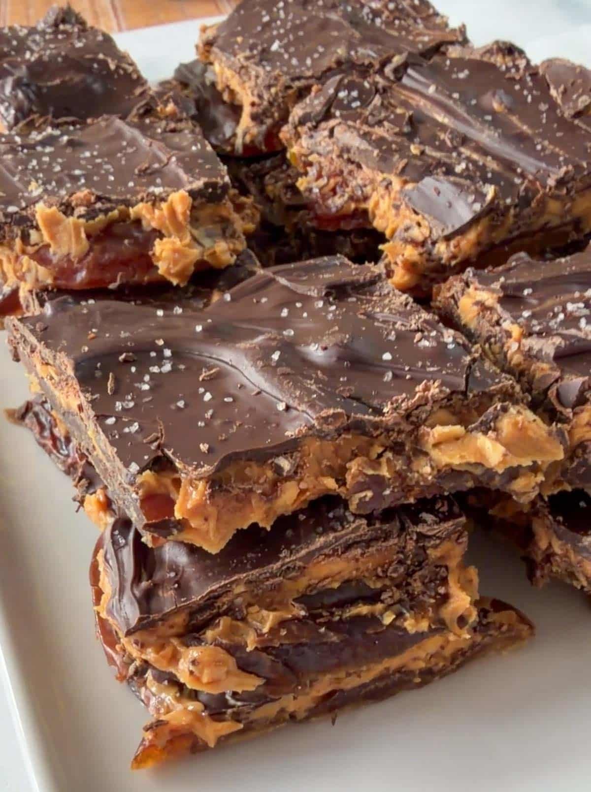 Date snickers stacked on a white plate, topped with sea salt and featuring a layered texture of chocolate, peanut butter, and crunchy ingredients.