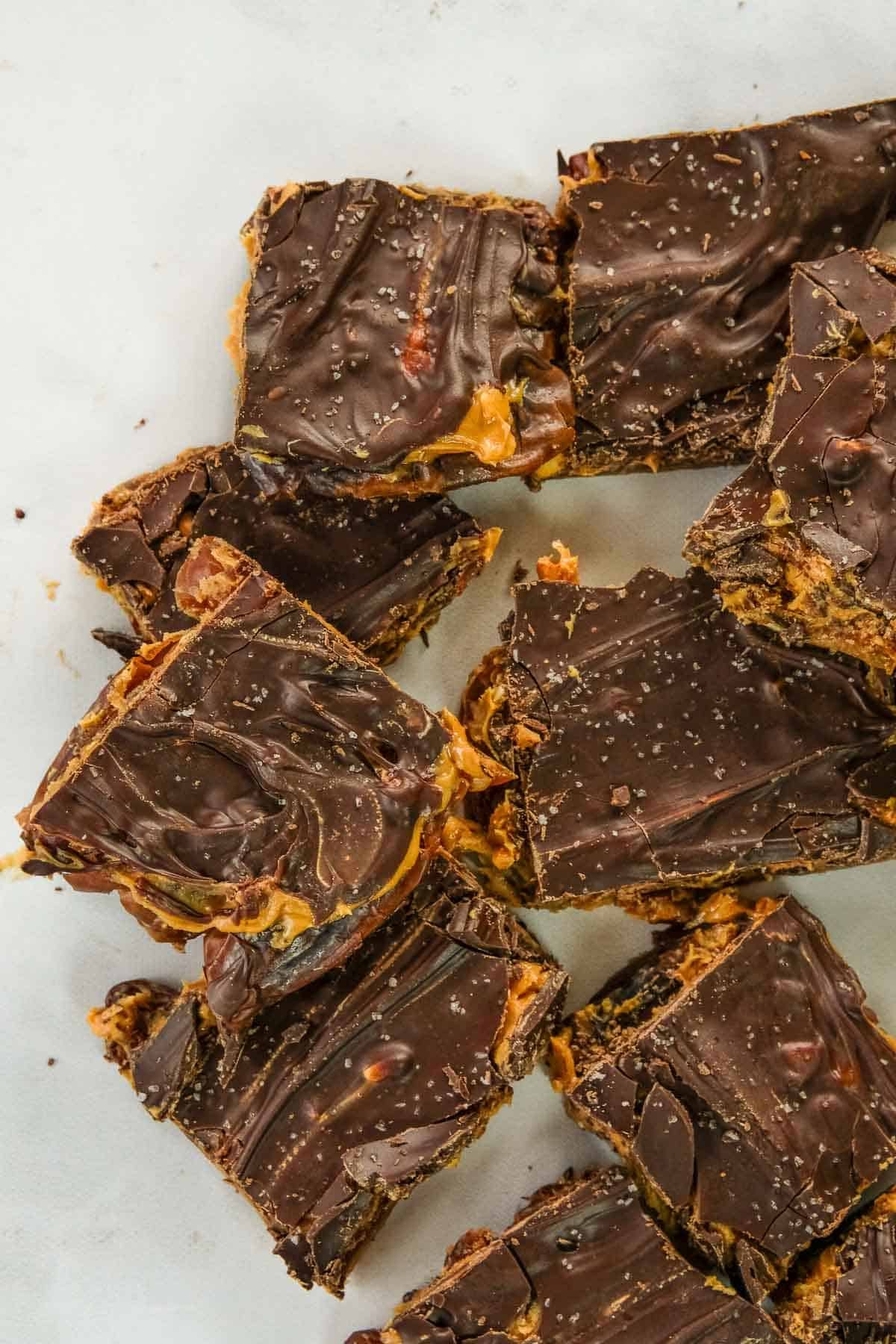 Squares of date snickers arranged on a white surface, with a slightly swirled and glossy chocolate topping sprinkled with sea salt.