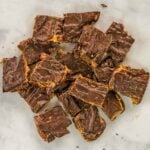 Squares of date snickers bark stacked on a white surface, with visible layers of chocolate and peanut butter and a swirl pattern on top.