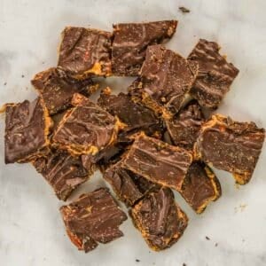 Squares of date snickers bark stacked on a white surface, with visible layers of chocolate and peanut butter and a swirl pattern on top.