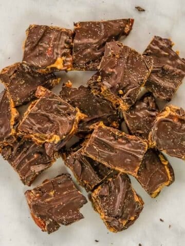 Squares of date snickers bark stacked on a white surface, with visible layers of chocolate and peanut butter and a swirl pattern on top.