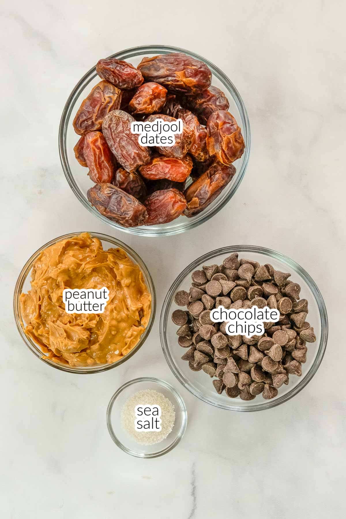 Three glass bowls on a white surface, containing ingredients for date snickers including medjool dates, chocolate chips, sea salt and peanut butter.