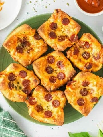 A green plate holds seven square pizza muffins with melted cheese.