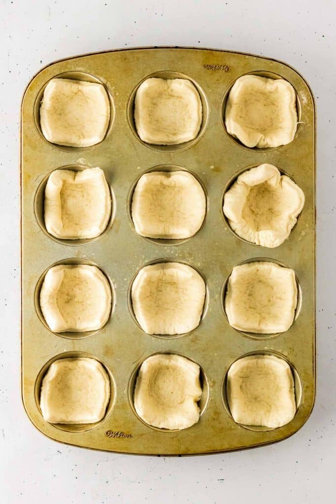 A standard muffin tin with twelve cavities, each lined with an uncooked piece of dough.