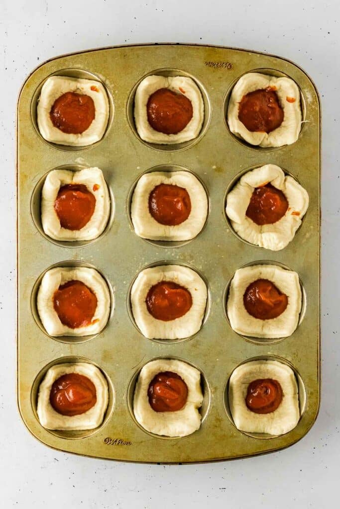 A muffin tin with twelve slices of dough inserted and filled with marinara sauce.