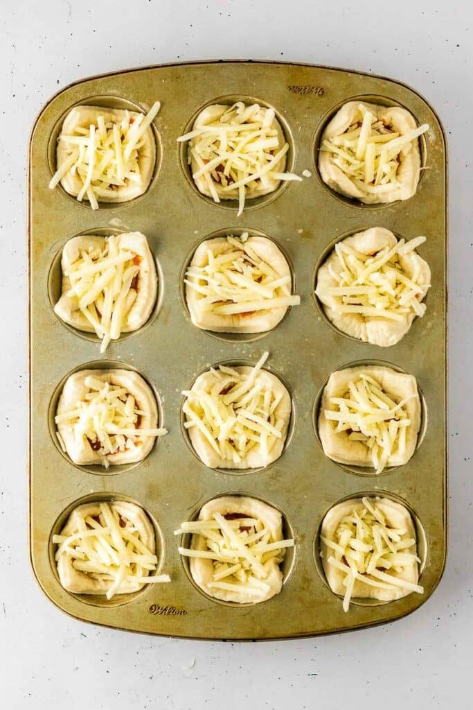 A muffin tin with twelve cups, each containing dough and sauce and topped with shredded cheese, ready to be baked.