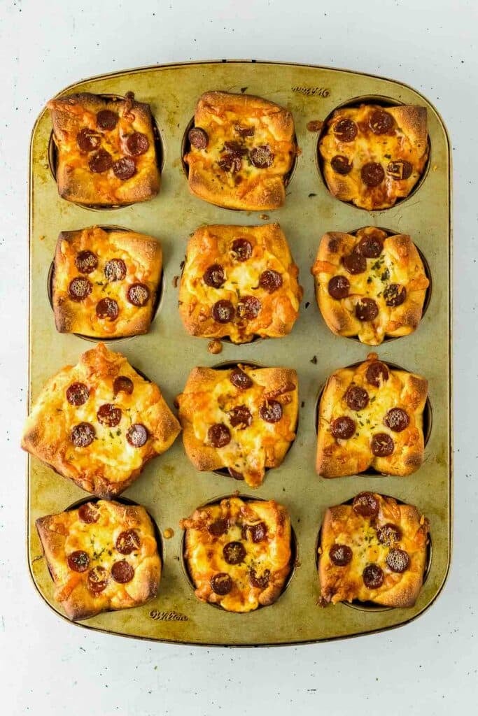 A muffin tin holding twelve baked pizza muffins with golden crusts, melted cheese, and slices of pepperoni on top.