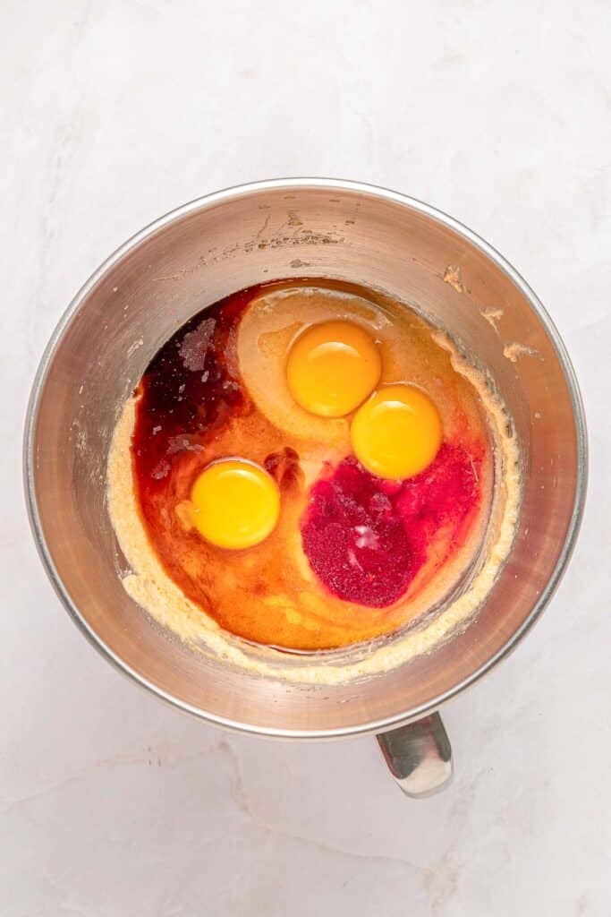 A metal mixing bowl containing flour, three raw eggs, red food coloring and vanilla extract.