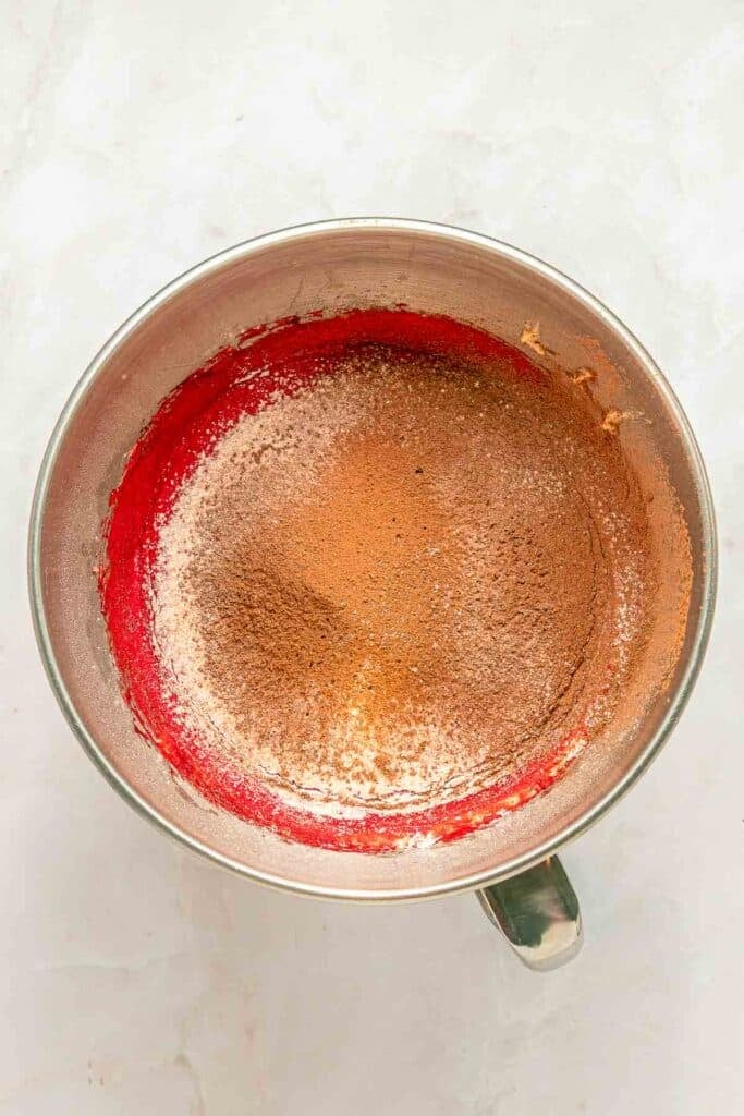 A metal mixing bowl containing flour and cocoa powder on top of a red batter, ready to be mixed.