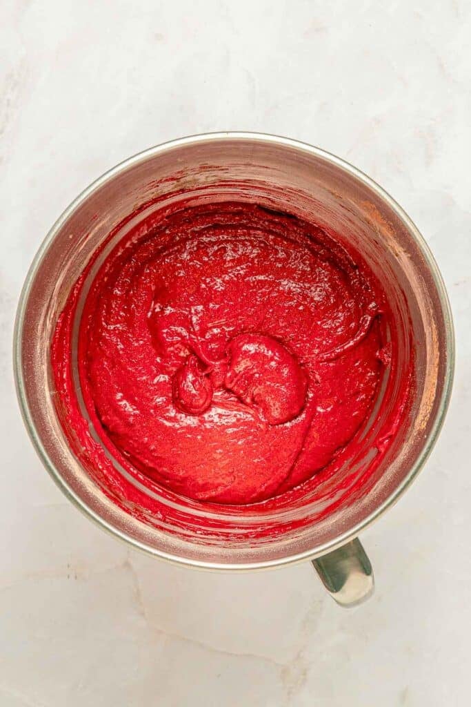 A mixing bowl filled with smooth, bright red cake batter on a light marble surface.