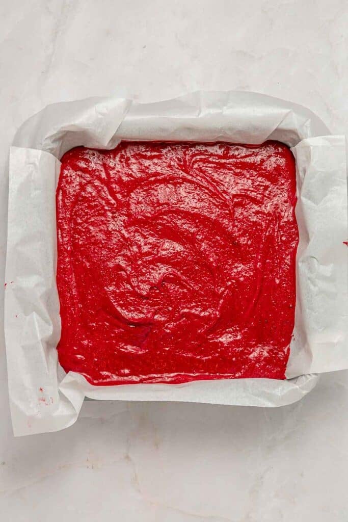 Square baking pan lined with parchment paper filled with raw red velvet cake batter on a light surface.