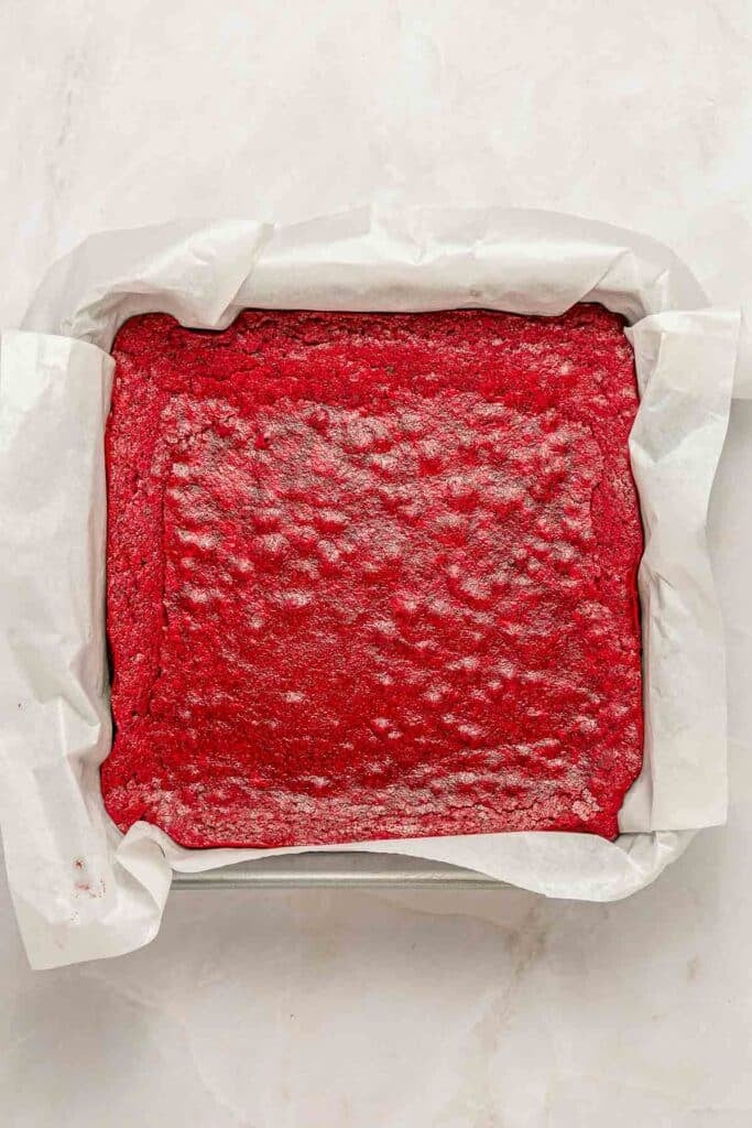 A square pan lined with parchment paper holds an uncut sheet of baked red velvet brownies on a light surface.