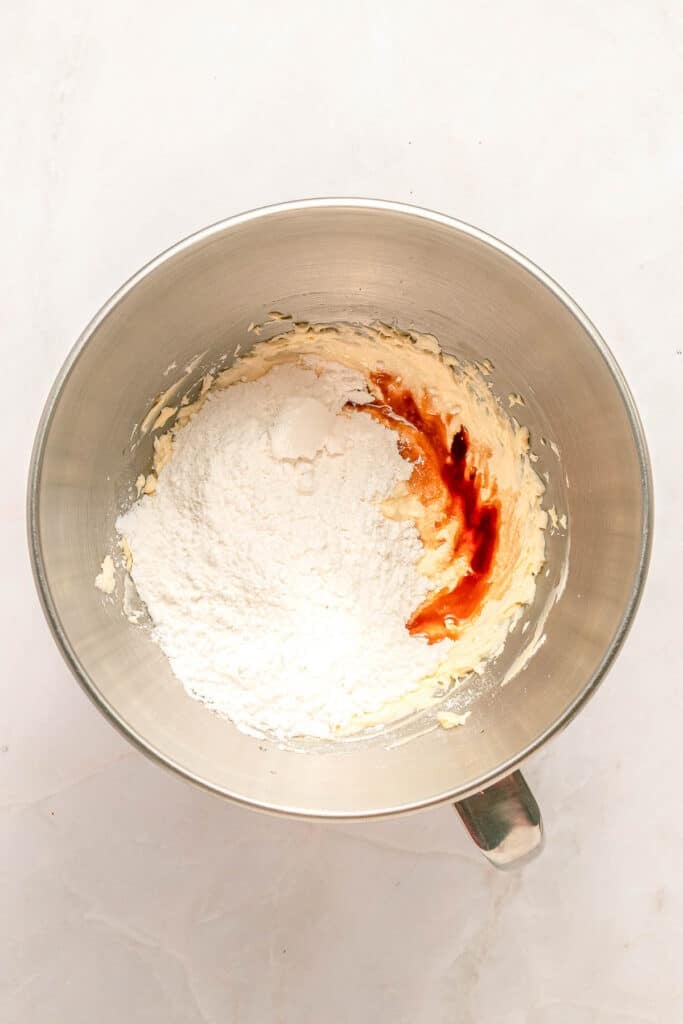 A metal mixing bowl with flour, vanilla extract, and creamed butter and sugar mixture on a white surface.