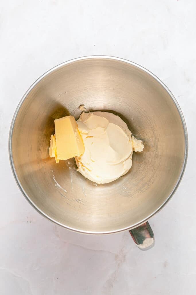 A metal mixing bowl contains a block of butter and cream cheese on a white surface.