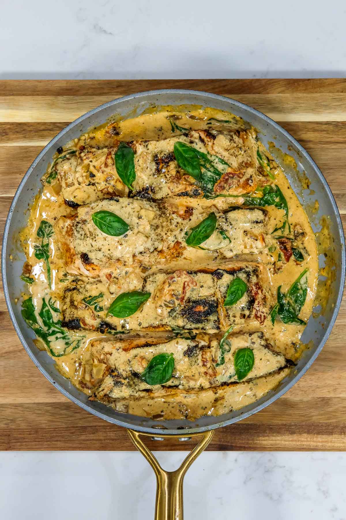 A skillet with cooked salmon fillets in a creamy sauce, garnished with fresh basil leaves, placed on a wooden cutting board.