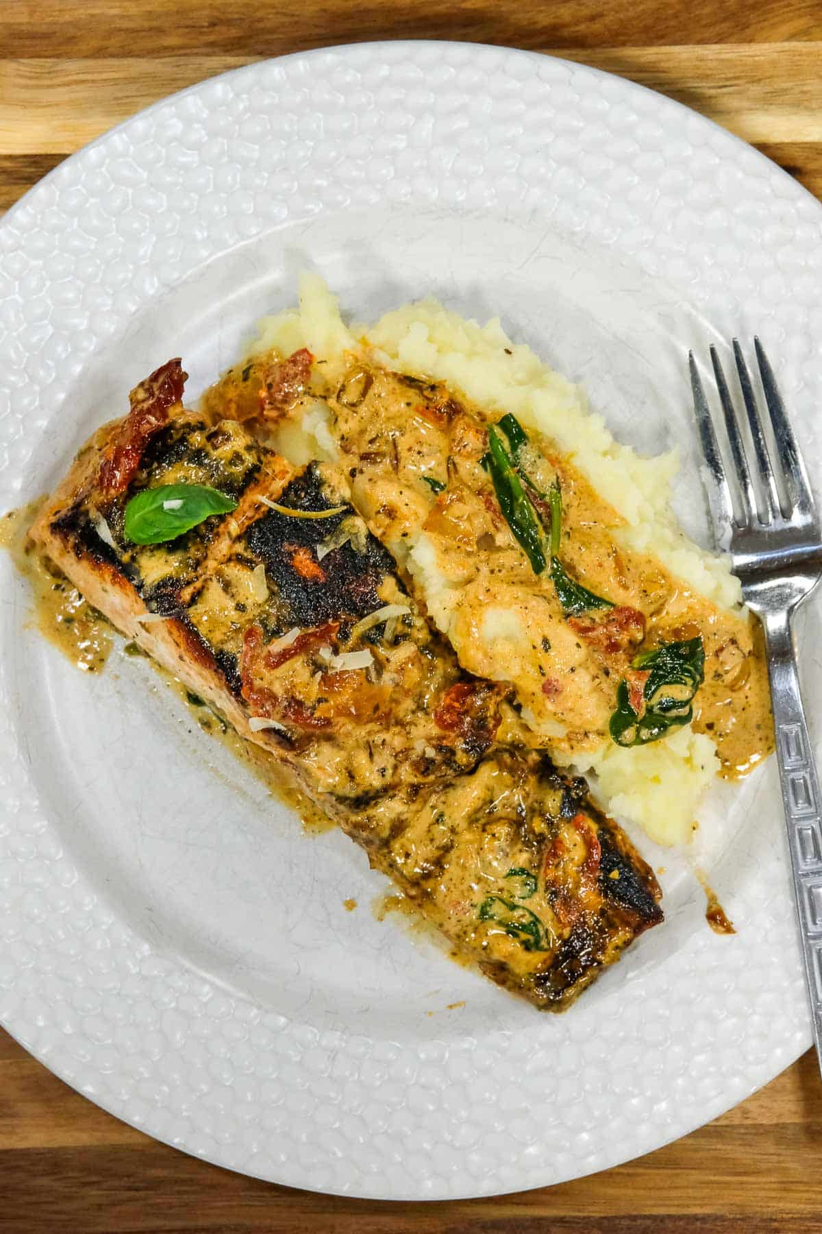 A piece of cooked tuscan salmon with creamy sauce and herbs, served with mashed potatoes on a white plate, with a fork on the side.