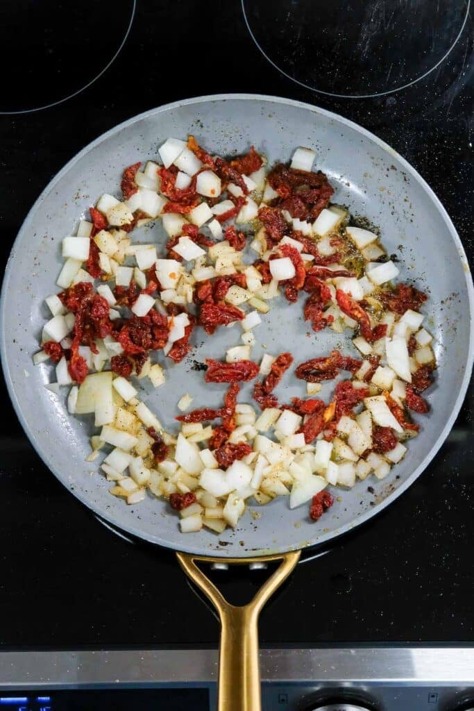 Diced onions and chopped sun-dried tomatoes are being sautéed in a frying pan on a stovetop.