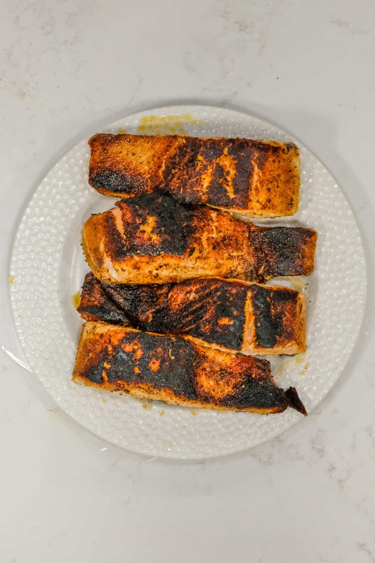 Four pieces of blackened, seasoned salmon are arranged in a row on a white textured plate, displayed on a light-colored surface.