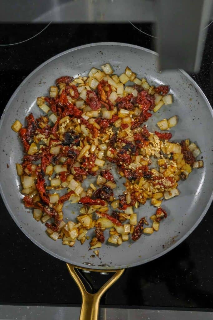 Diced onions, sun-dried tomatoes, and minced garlic sautéing in a gray skillet on a stovetop.
