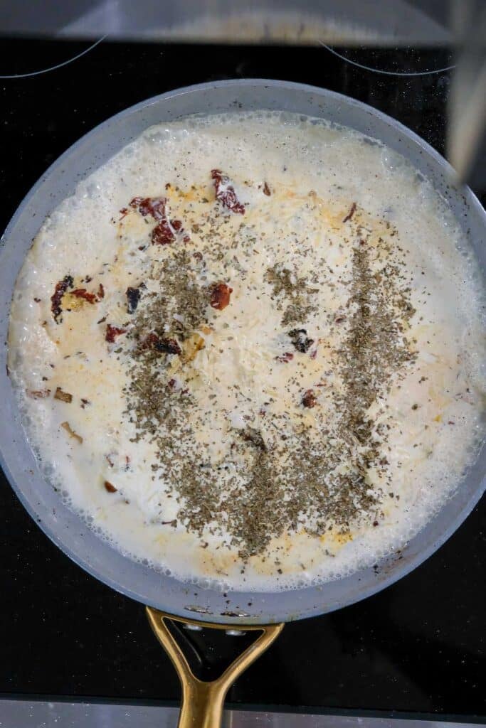 A frying pan on a stove containing cream, herbs, and pieces of sun-dried tomato being cooked together.
