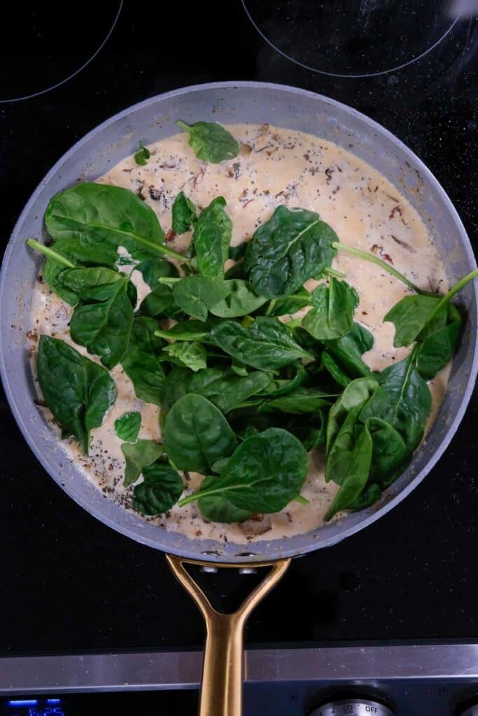A pan on a stovetop contains creamy sauce with herbs, pieces of sun-dried tomato, and fresh spinach leaves on top, not yet stirred in.