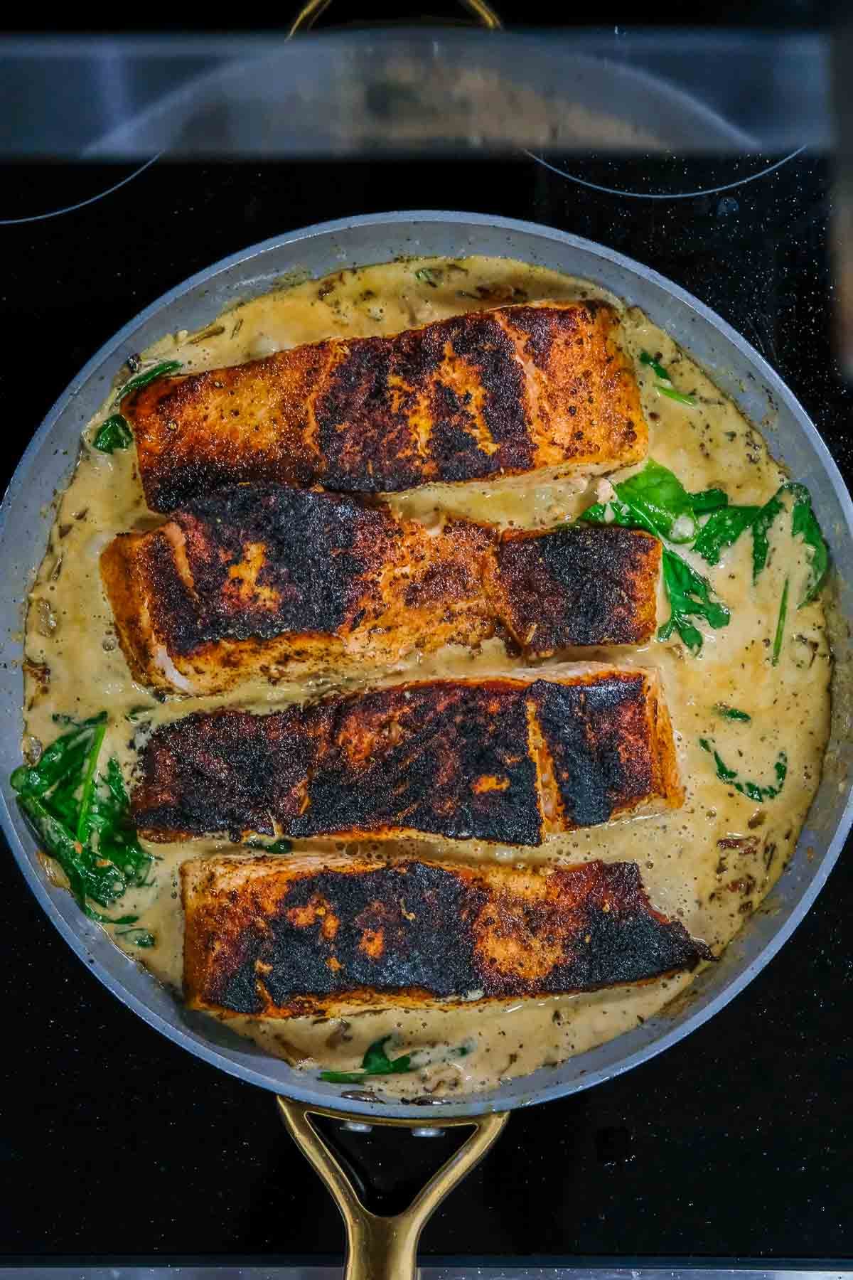 Four blackened salmon fillets are cooking in a creamy spinach sauce in a large skillet on a stovetop.