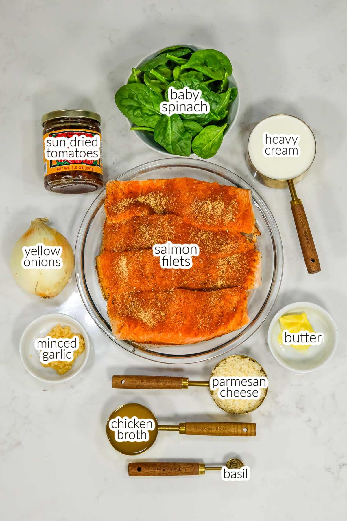 Overhead view of ingredients for Tuscan Salmon including seasoned salmon filets surrounded by small bowls of baby spinach, heavy cream, sun-dried tomatoes, yellow onions, minced garlic, chicken broth, butter, parmesan cheese, and basil.