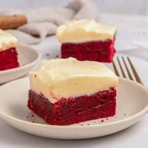 A square piece of red velvet cake with cream cheese frosting sits on a white plate, with another piece and a fork in the background.