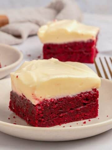 A square piece of red velvet cake with cream cheese frosting sits on a white plate, with another piece and a fork in the background.