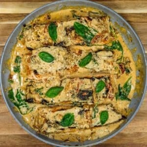 Tuscan salmon fillets in a creamy sauce with spinach, sun-dried tomatoes, and basil leaves in a skillet, viewed from above.