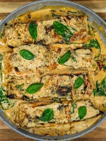 Tuscan salmon fillets in a creamy sauce with spinach, sun-dried tomatoes, and basil leaves in a skillet, viewed from above.