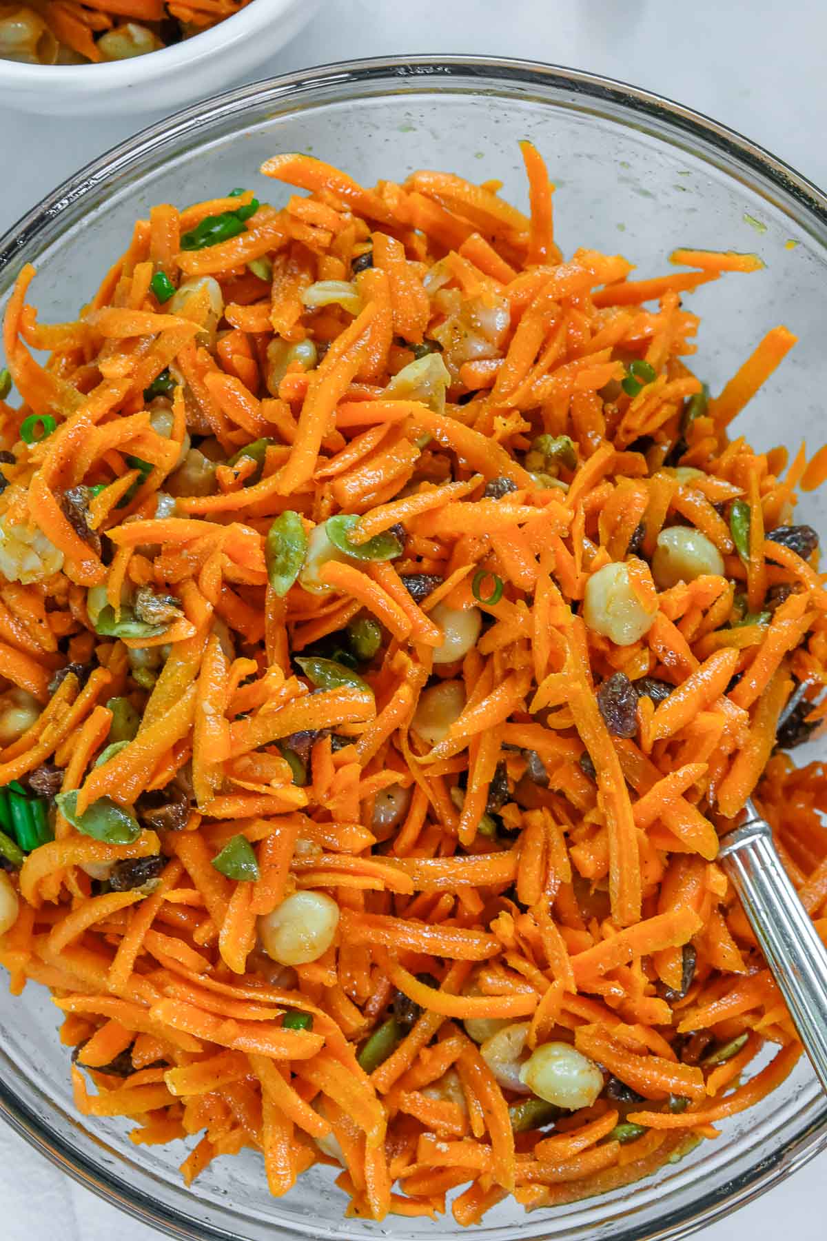 A glass bowl filled with carrot salad mixed with chickpeas, green onions, and nuts, with a fork resting inside.