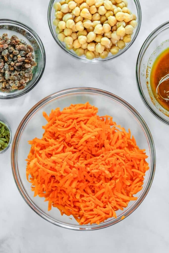 Four glass bowls on a white surface: one with shredded carrots, one with chickpeas, one with raisins, and one with dressing. Partial view of a bowl with pumpkin seeds.