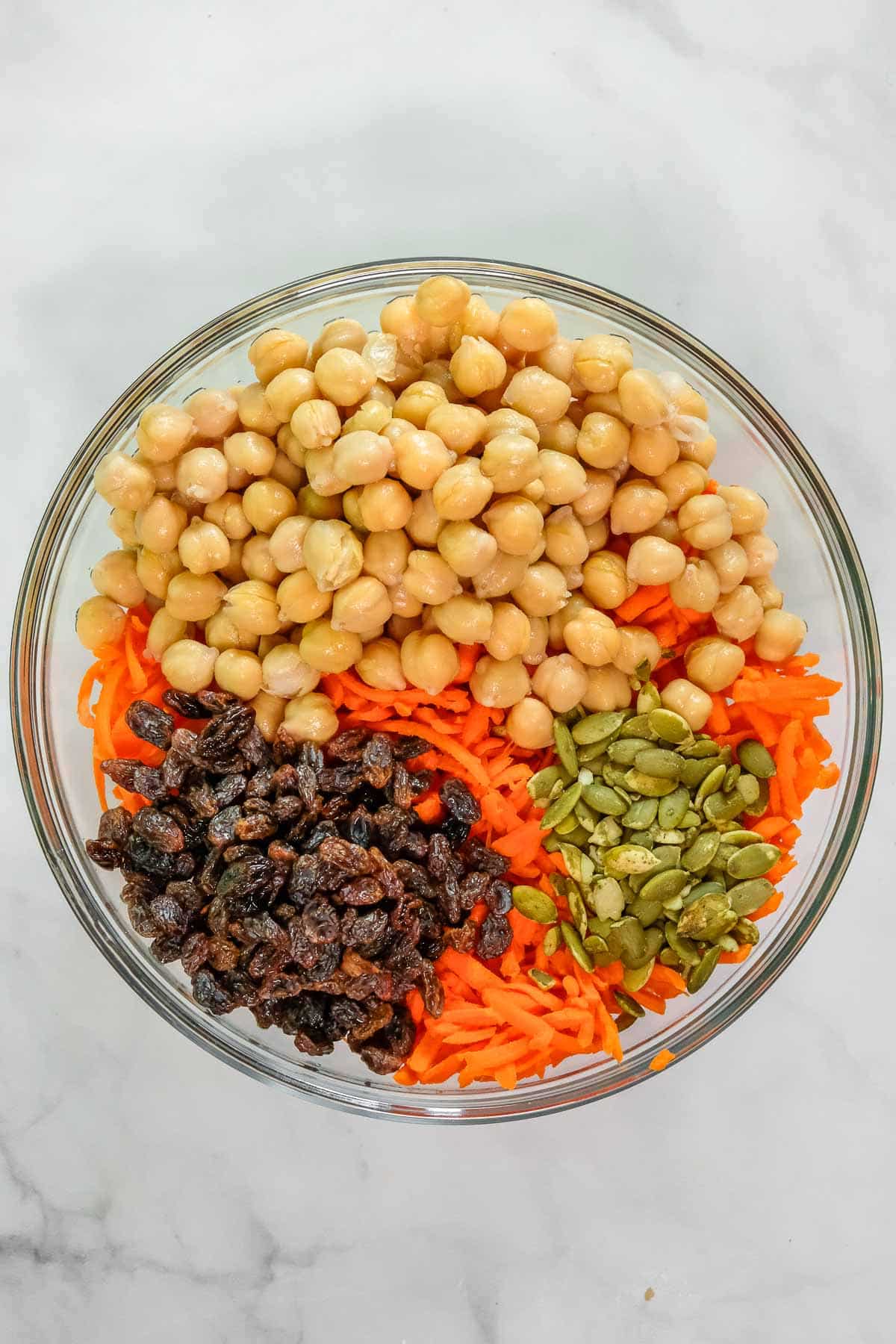 A glass bowl containing chickpeas, shredded carrots, raisins, and pumpkin seeds arranged in separate sections on a light surface.