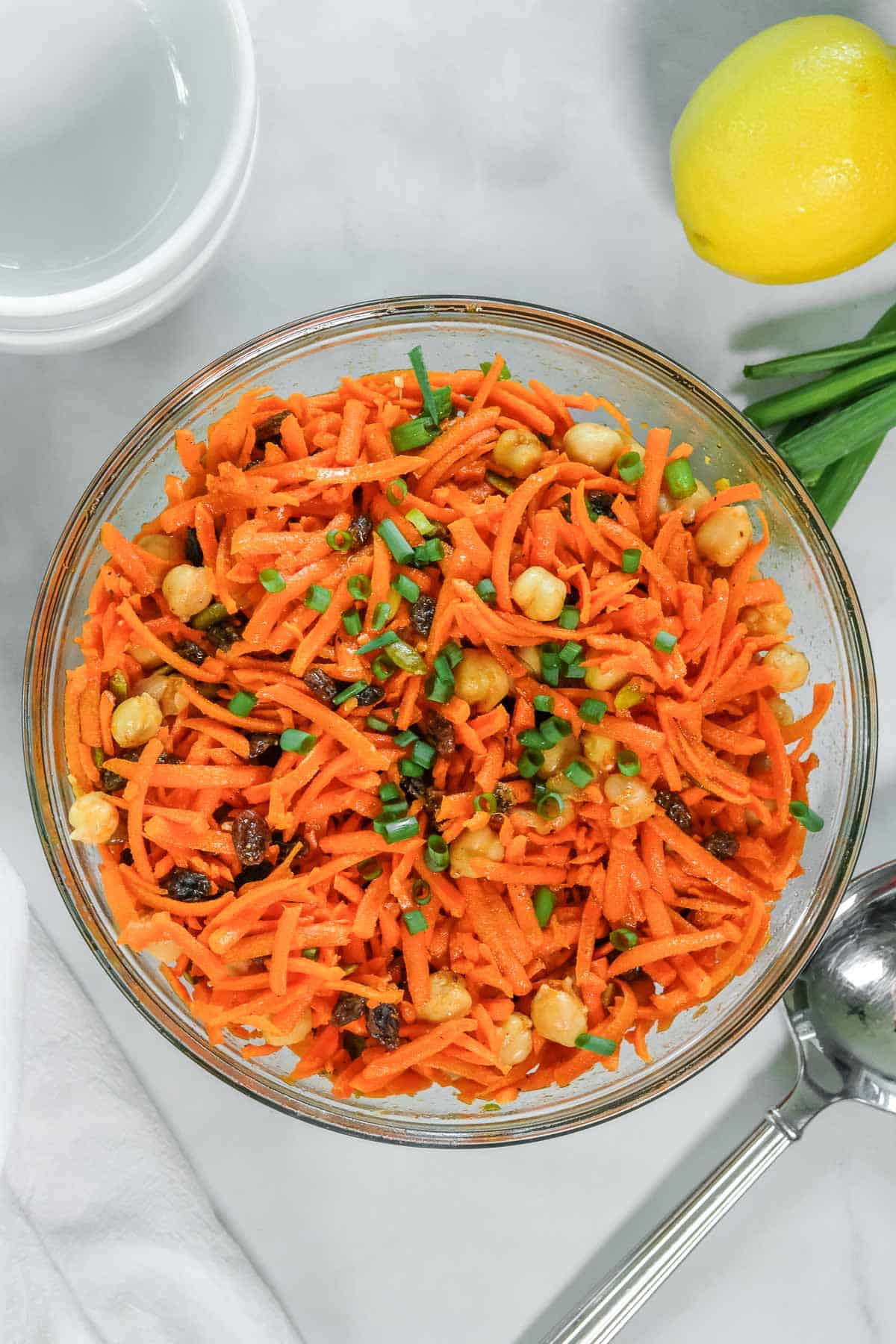 A glass bowl filled with shredded carrot salad mixed with chickpeas, raisins, and chopped green herbs, placed on a white surface next to a lemon and some green onions.