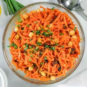 A glass bowl filled with carrot salad mixed with chickpeas, raisins, and chopped green onions.