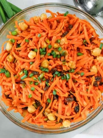 A glass bowl filled with carrot salad mixed with chickpeas, raisins, and chopped green onions.