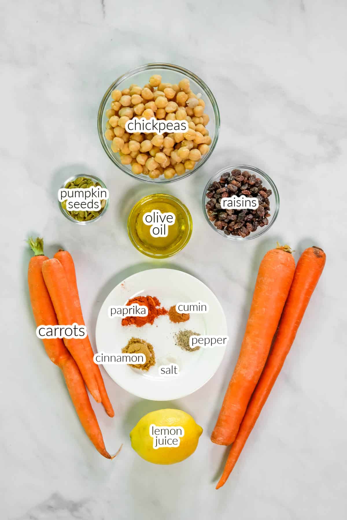 Ingredients for carrot salad laid out including Carrots, chickpeas, pumpkin seeds, raisins, olive oil, lemon juice, and various spices including paprika, cumin, cinnamon, salt, and pepper are arranged on a white surface.