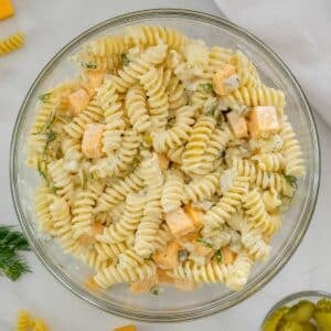 A glass bowl filled with Dill Pickle Pasta Salad mixed with creamy dressing, diced cheese cubes, chopped dill, and small pieces of pickle.