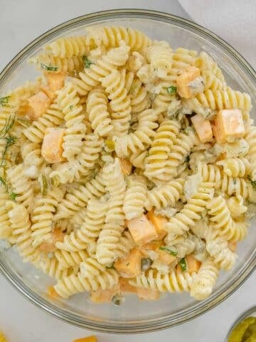 A glass bowl filled with Dill Pickle Pasta Salad mixed with creamy dressing, diced cheese cubes, chopped dill, and small pieces of pickle.