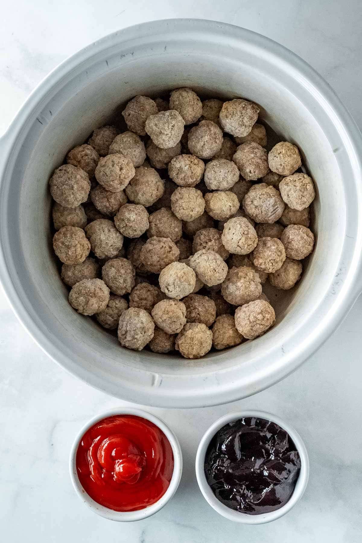 A slow cooker filled with frozen meatballs, with a bowl of ketchup and a bowl of grape jelly placed beside it on a white countertop.