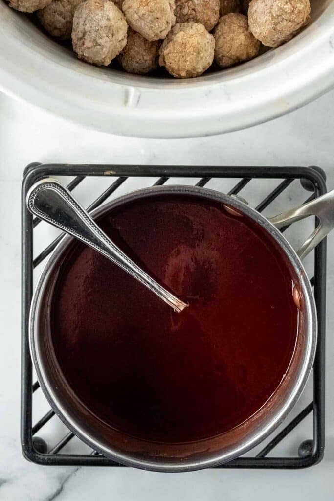 A saucepan filled with grape jelly and ketchup sauce and a spoon sits on a trivet; a slow cooker with uncooked meatballs is partially visible above.