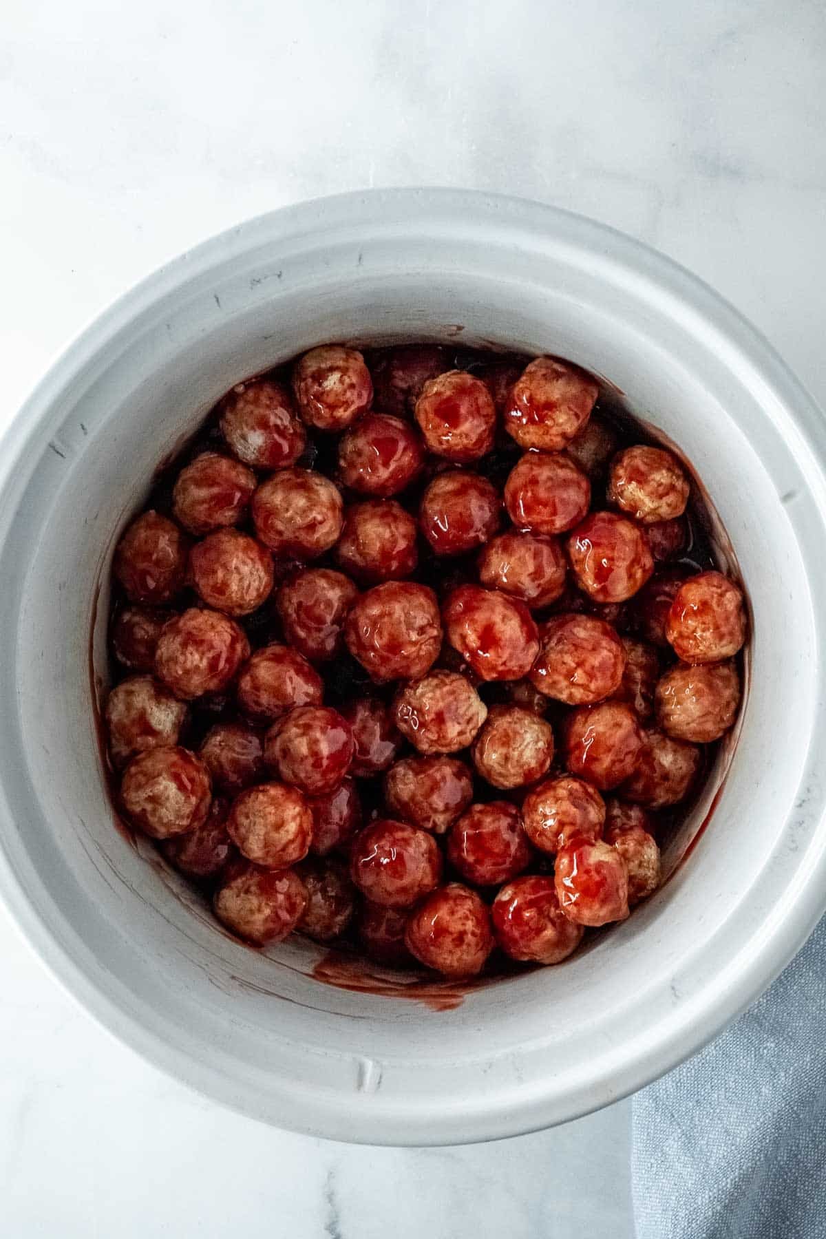 A white slow cooker filled with meatballs coated in a grape jelly sauce, viewed from above on a marble