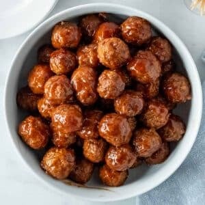 A white bowl filled with grape jelly and ketchup meatballs on a white marble countertop.